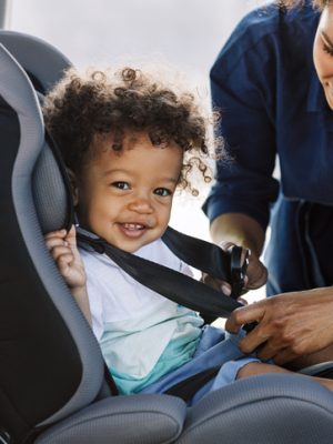 Side view of a happy little boy looking at camera while his mother buckling him in a car seat